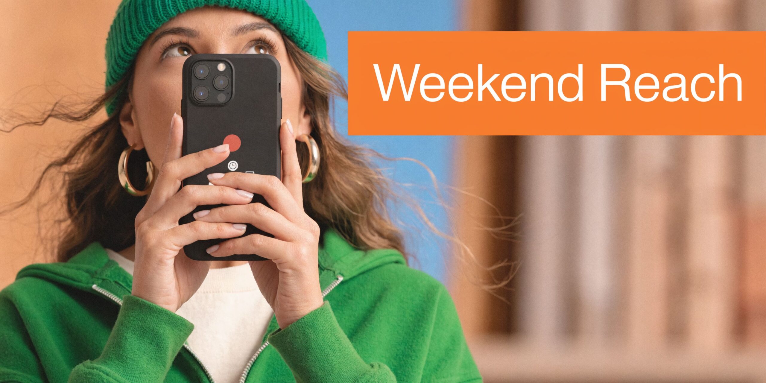 A woman wearing a green beanie and hoodie holding a black smartphone to her face while smiling.