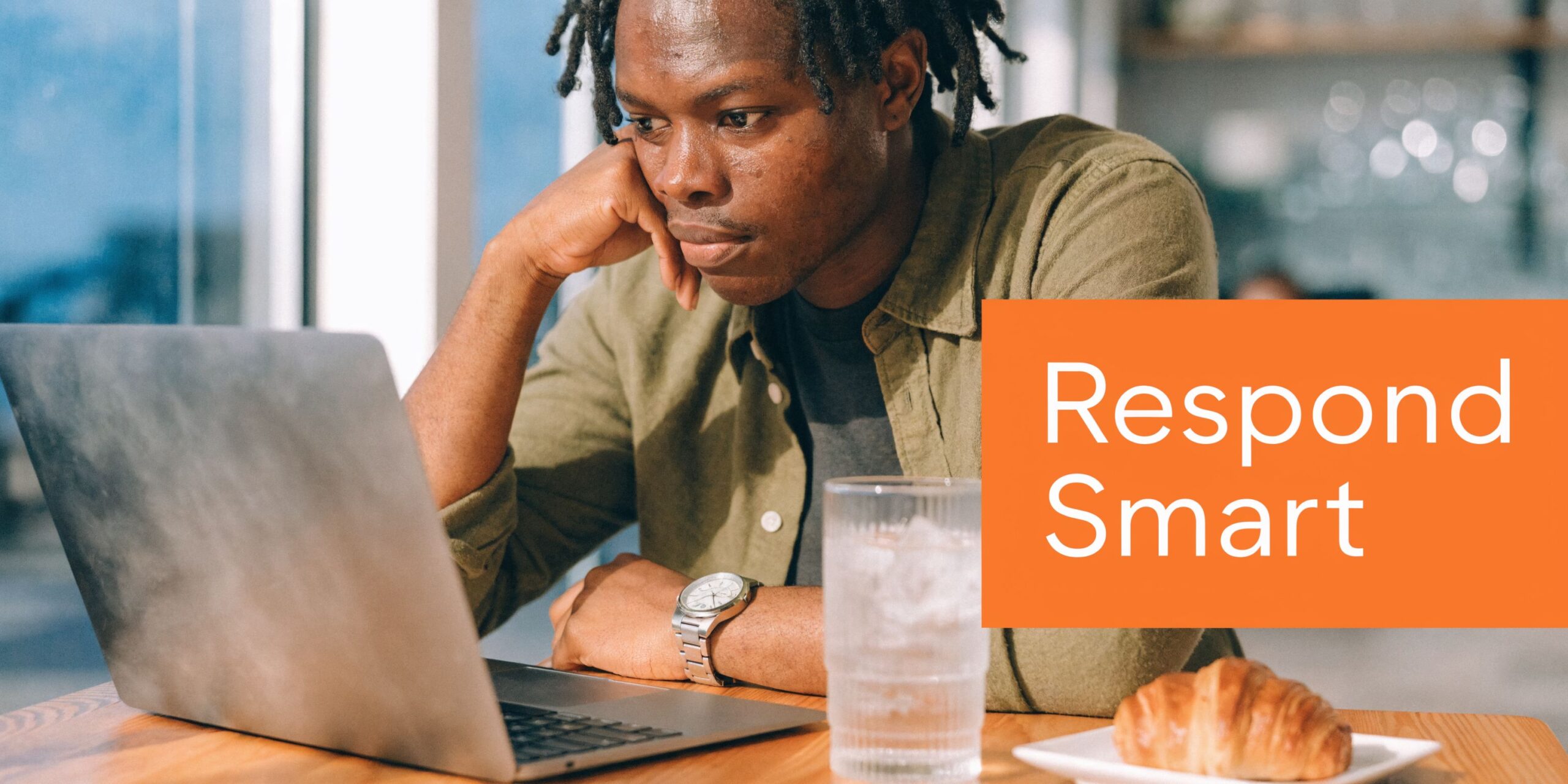 A young man with dreadlocks working on his laptop while sitting at a table with a croissant.