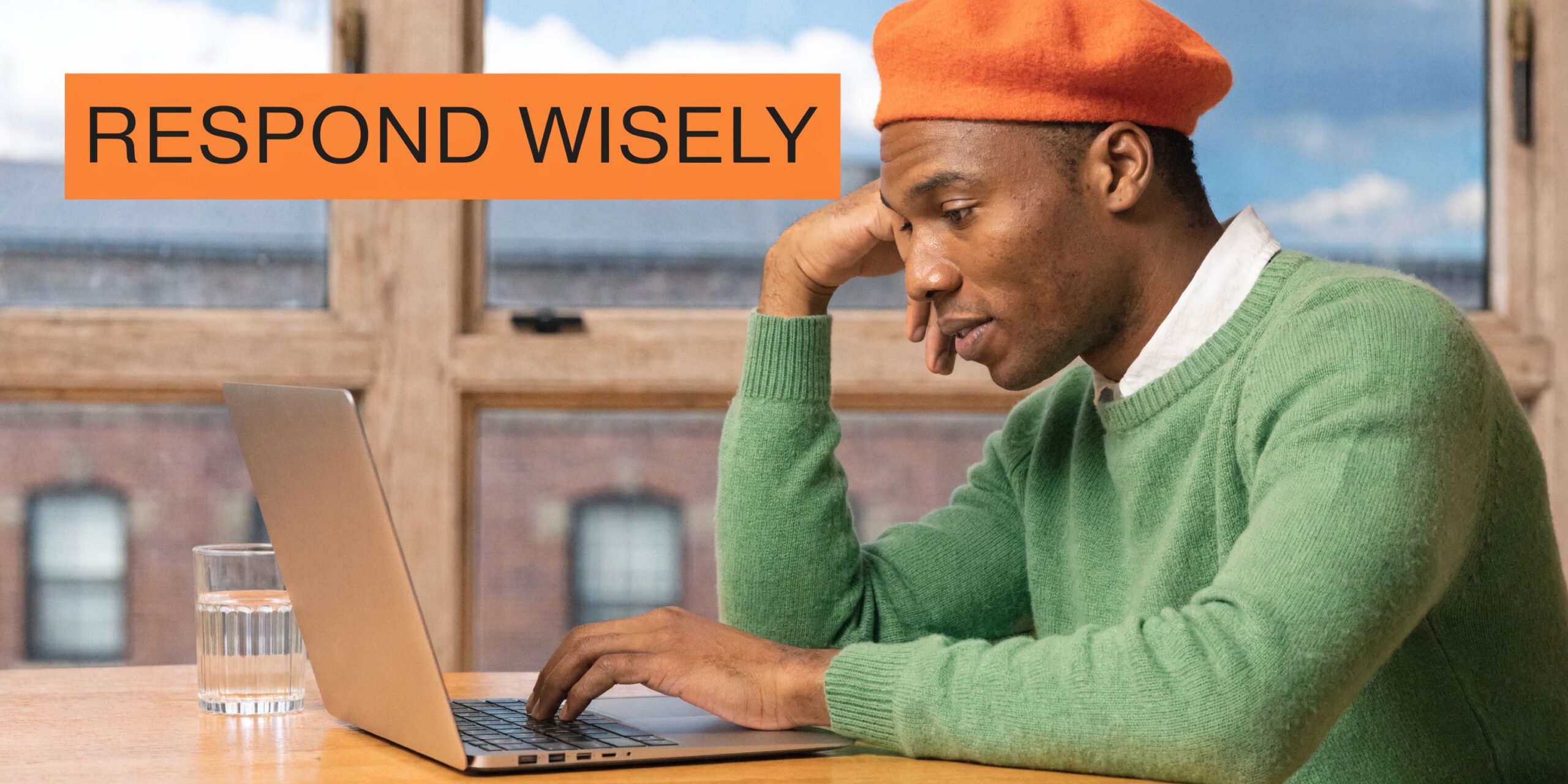 A man wearing an orange beret and green sweater types on a laptop while working from home.